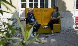 Auf dem gelben Zuhörbänkli sitzen zwei Frauen und sprechen miteinander.
