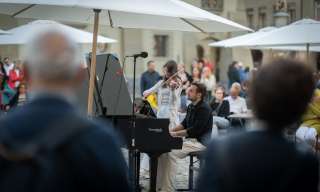 Ein Mann sitzt an einem Flügel und eine Frau spielt daneben Geige. Das Publikum steht um den Flügel herum und singt mit. Foto: BernCityPiano
